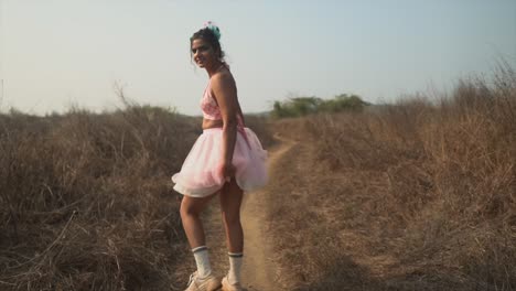 the back of a happy carefree asian female walking along a path in a dry grass field wearing a pink tutu, turning around playfully holding her dress and smiling, india