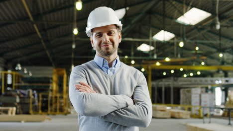 trabajador caucásico con casco y posando para la cámara con los brazos cruzados en una fábrica