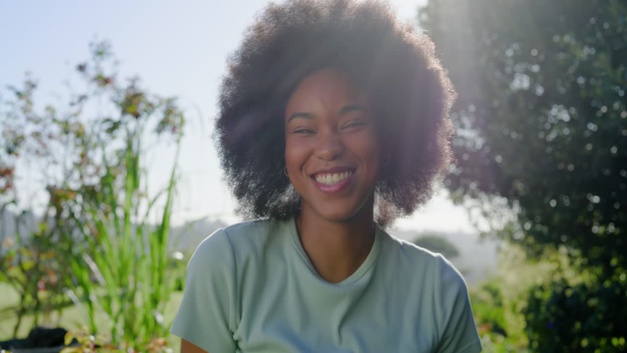 African American woman standing in garden in green tee, laughing, adjusting hair to sun flare