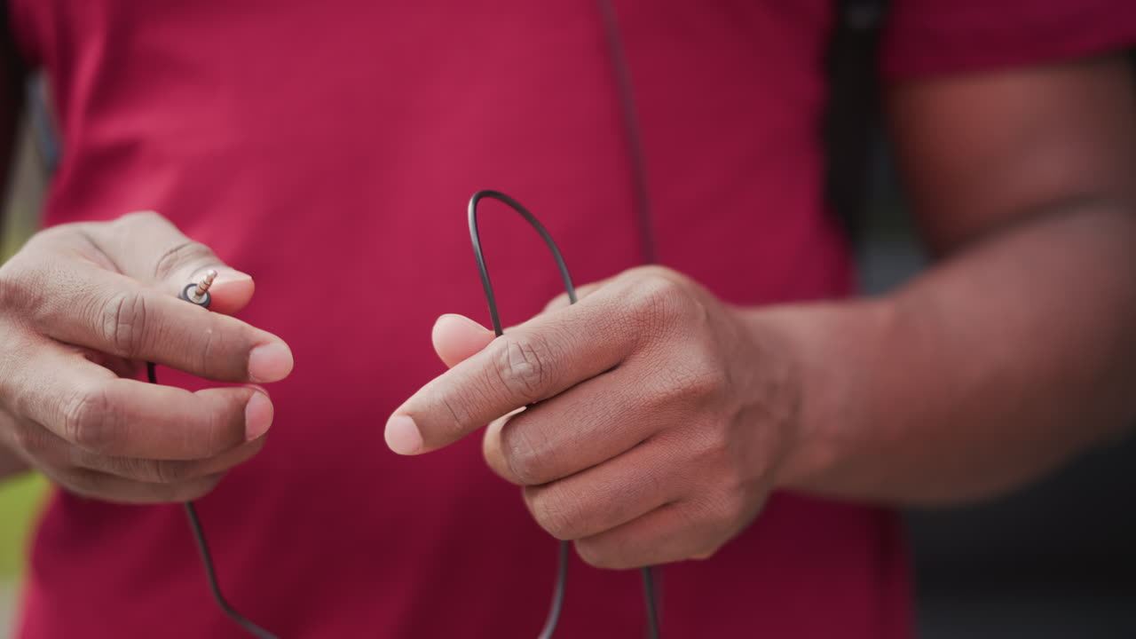 black street musician untangles worn earphones, closeup of dusty hands in red shirt manipulating tangled audio cable, careful fingers sorting cord and plug, outdoor urban sidewalk mood, preparation