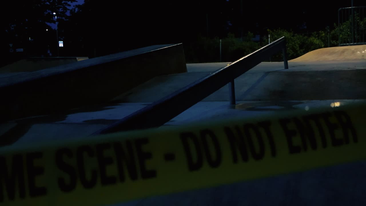 Close up or closeup footage of a yellow colored police Crime Scene Do Not Enter tape caution tape ribbon pulled in front of a concrete gray evening or night skatepark with bokeh street light at back