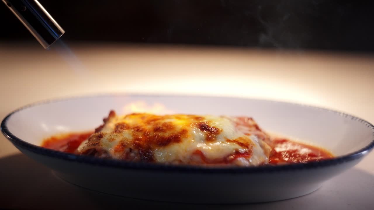 Close up view in slow motion of a torch flaming the cheese of a lasagna in a fine dining restaurant counter.