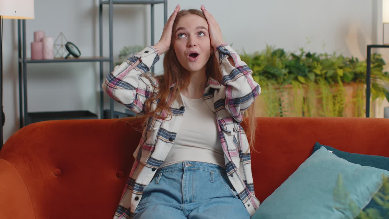 흥분한 젊은 여성은 소파에서 집에서 갑작스러운 승리에 충격을 받아 놀라서 손을 들어 올렸습니다.