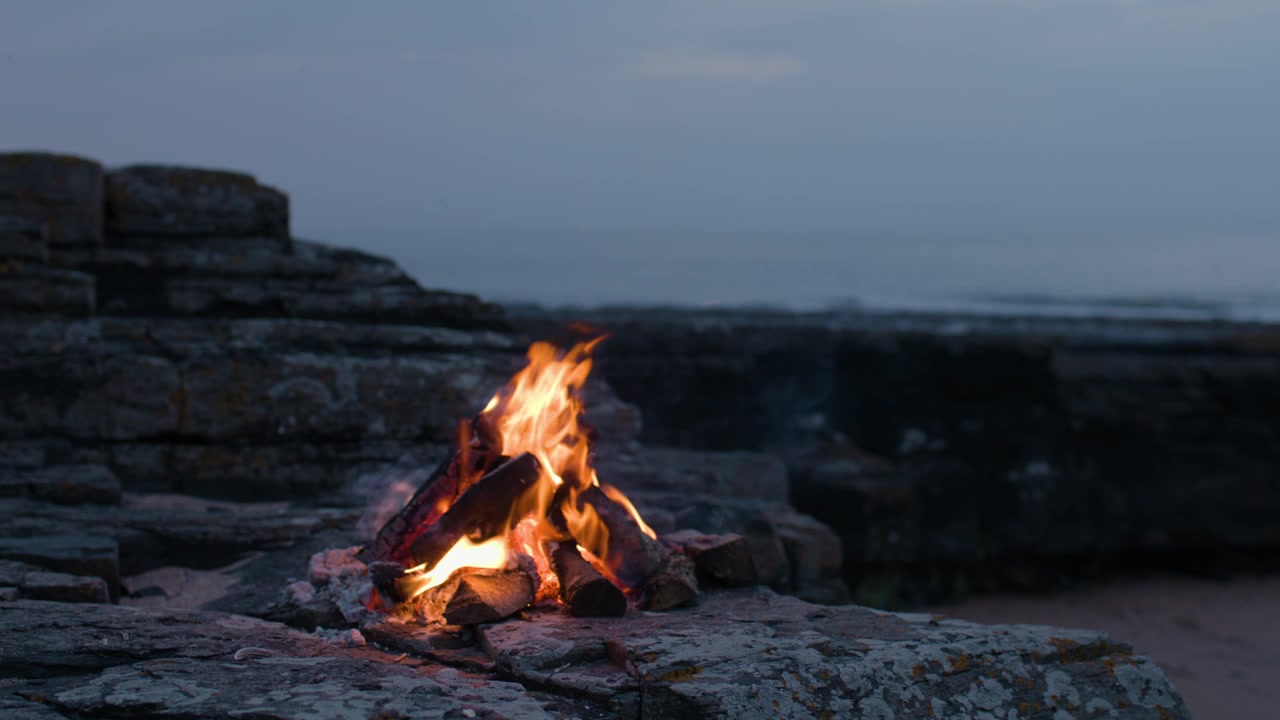 Un falò ardente brucia su una costa rocciosa e frastagliata nella luce soffusa del tardo pomeriggio, con una leggera brezza marina che occasionalmente muove le fiamme contro uno sfondo di oceano calmo.