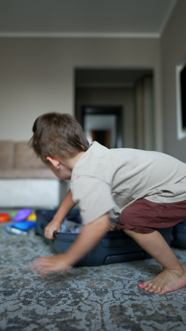 Kid appears out of the big grey suitcase lying on the floor. Baby boy stands up and tosses his clothes up. Vertical video.