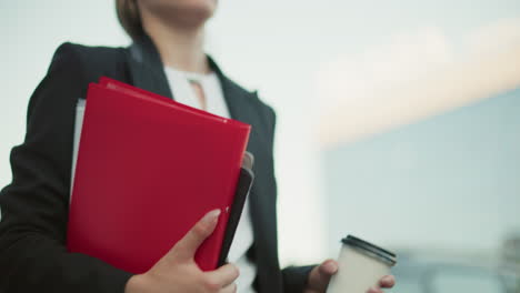 close up of banker in black suit holding file folder and coffee cup while walking along path with blurred background of parked cars and urban buildings during bright morning commute to office
