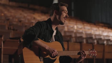 a handsome guy plays an acoustic guitar in an abandoned cinema. the musician sings a song and accompanies on the guitar