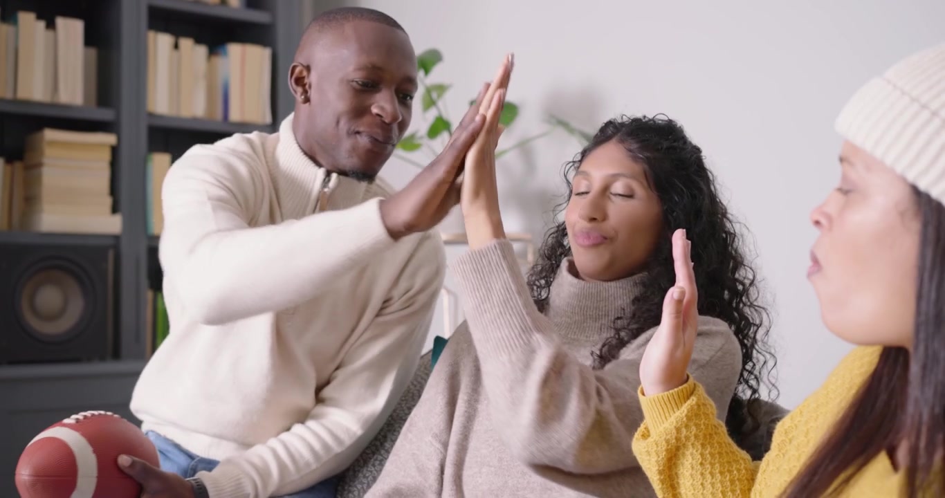 Afro-Amerikaanse vrienden reageren op een scherm op de bank, met een kom, pretzels en een voetbal, terwijl ze elkaar een high five geven.