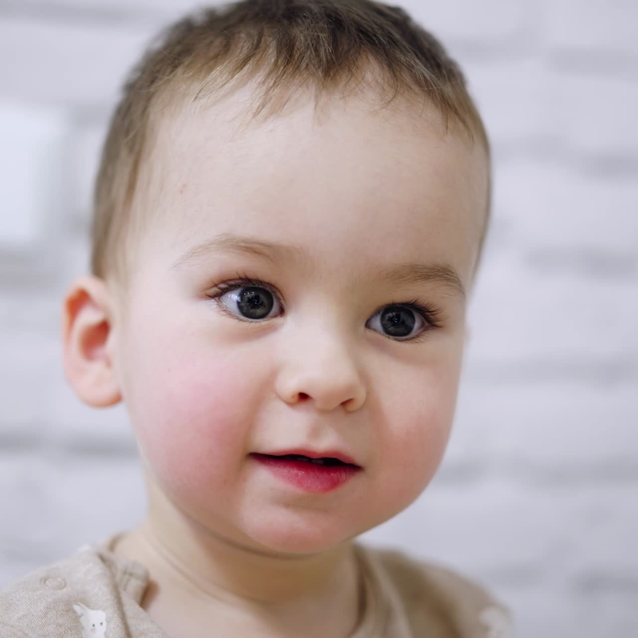 Adorable big-eyed baby boy with mousy hair. Portrait of a calm Caucasian kid focused on something. Close up. Blurred white backdrop