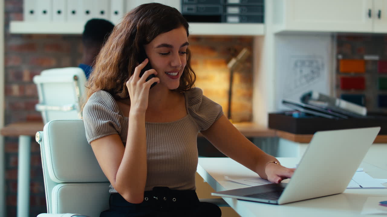 joven mujer de negocios sonriente trabajando en una computadora portátil en el escritorio en la oficina hablando en el teléfono móvil