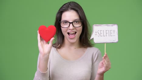 young happy beautiful woman holding red heart and selfie paper sign