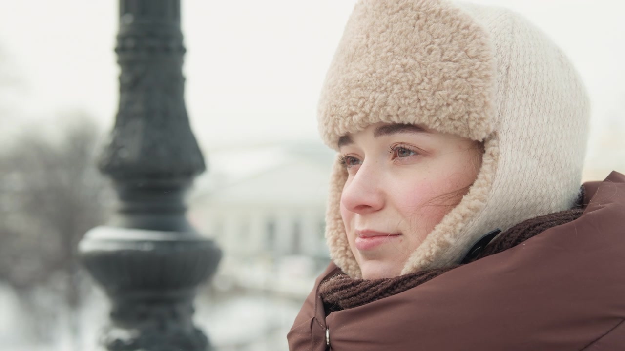 Giovane donna con cappello da trapper foderato di pelliccia e giacca invernale imbottita si sistema gli occhiali e il cappello su un marciapiede urbano innevato, con uno sguardo laterale calmo, un sorriso dolce, uno sfondo cittadino sfocato e una luce stagionale fredda.