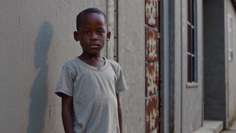 serious young african child standing still against weathered gray wall, gazing directly with profound vulnerability and silent resilience in remote village setting
