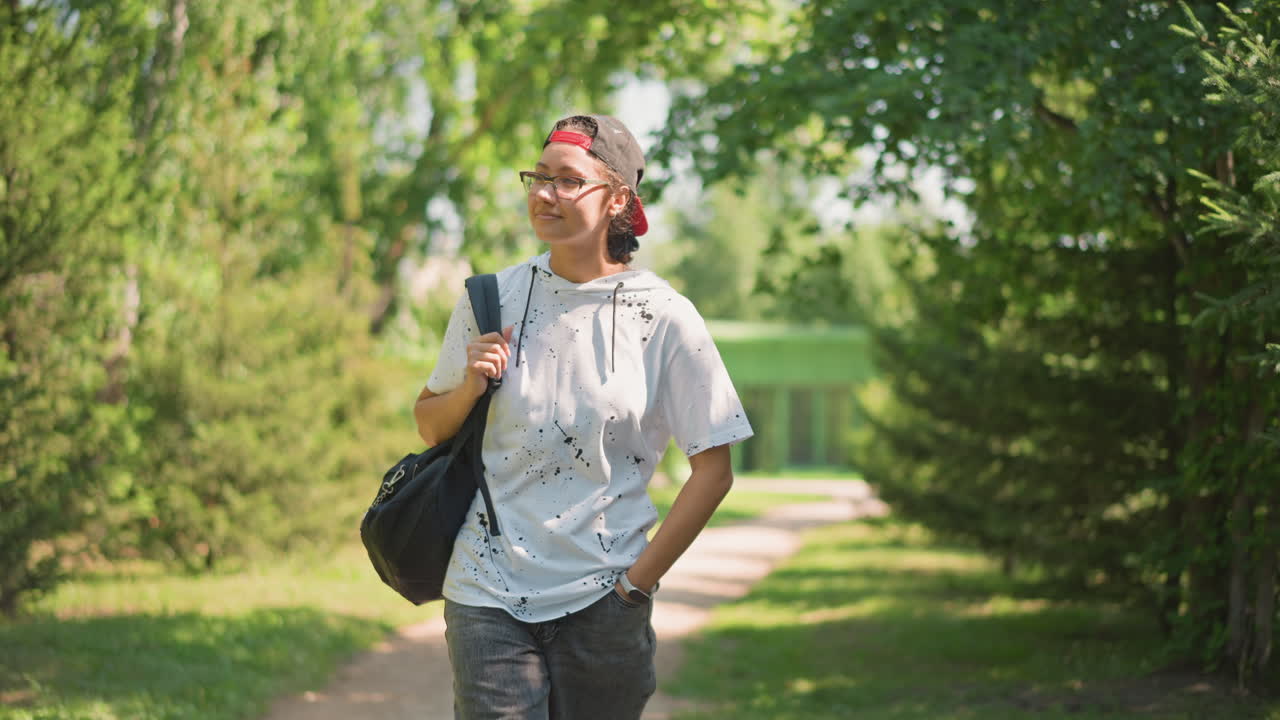 정면 프로필, 미소 짓는 학생이 어깨 가방을 메고 손을 주머니에 넣은 채 빨간 모자와 안경을 쓰고 초록색 공원 길을 따라 걷고 있어요. 배경에는 다리가 보이고, 밝은 햇빛 아래 자신감 넘치는 발걸음으로 여유로운 분위기를 풍기고 있습니다.