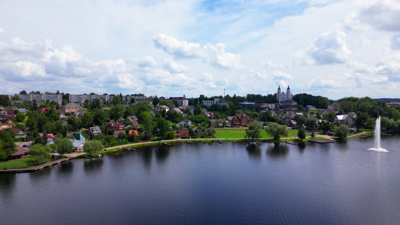 Wide aerial view of Zarasai town with lake and water fountain, historic church on the hill, and residential buildings nestled in forest. Shot in Zarasai, Lithuania (Zarasai, Lietuva)