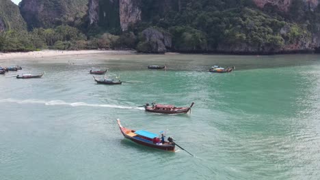 aerial view of longtail boat leaving beach with pan reveal of limestone cliffs in background at railay, thailand