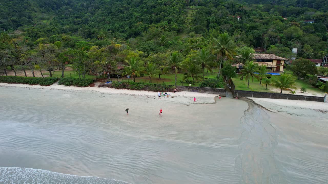 toma aérea en órbita de niños jugando en la playa con un bosque verde con montañas en el fondo y un hermoso mar con suaves olas rompiendo