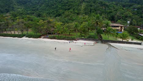 Toma-Aérea-En-órbita-De-Niños-Jugando-En-La-Playa-Con-Un-Bosque-Verde-Con-Montañas-En-El-Fondo-Y-Un-Hermoso-Mar-Con-Suaves-Olas-Rompiendo