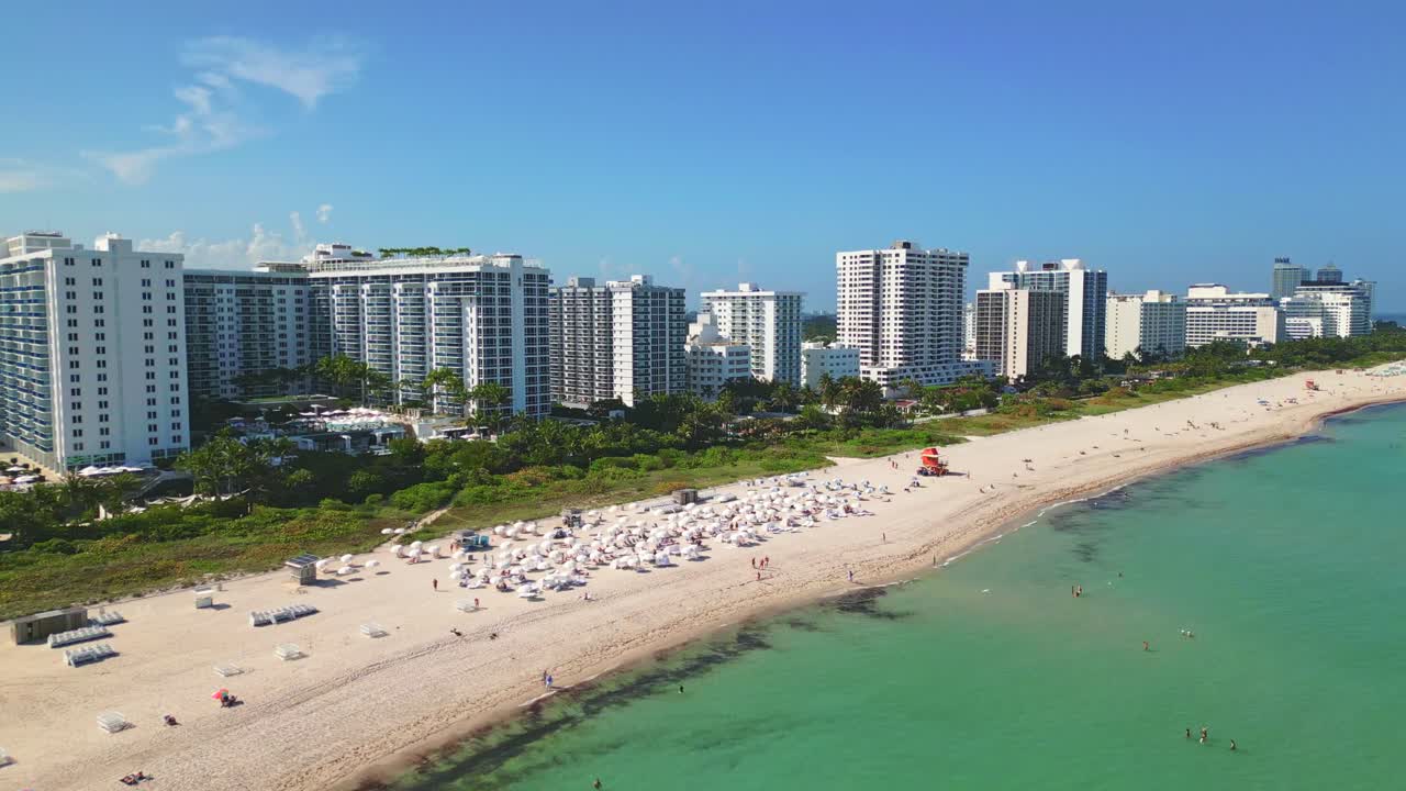 4K Drone glides over South Beach, Miami, moving from the shore into the turquoise ocean, unveiling the stunning coastline lined with white sands, swaying palms, and a row of luxury resort hotels.