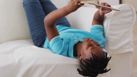 niño afroamericano sonriendo mientras usa una tableta digital mientras está acostado en el sofá en casa