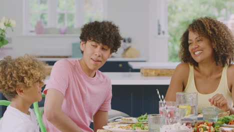 Multi-Racial-Family-Sitting-Around-Table-In-Kitchen-At-Home-Eating-Meal-Together