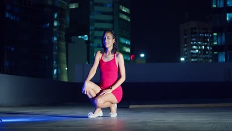 a young girl in a red dress stands on a rooftop at night, with city buildings silhouetted against the dark sky behind her