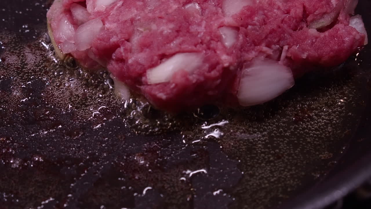 primer plano: hamburguesa de carne molida rosa cruda y cebollas chisporroteando en una sartén caliente