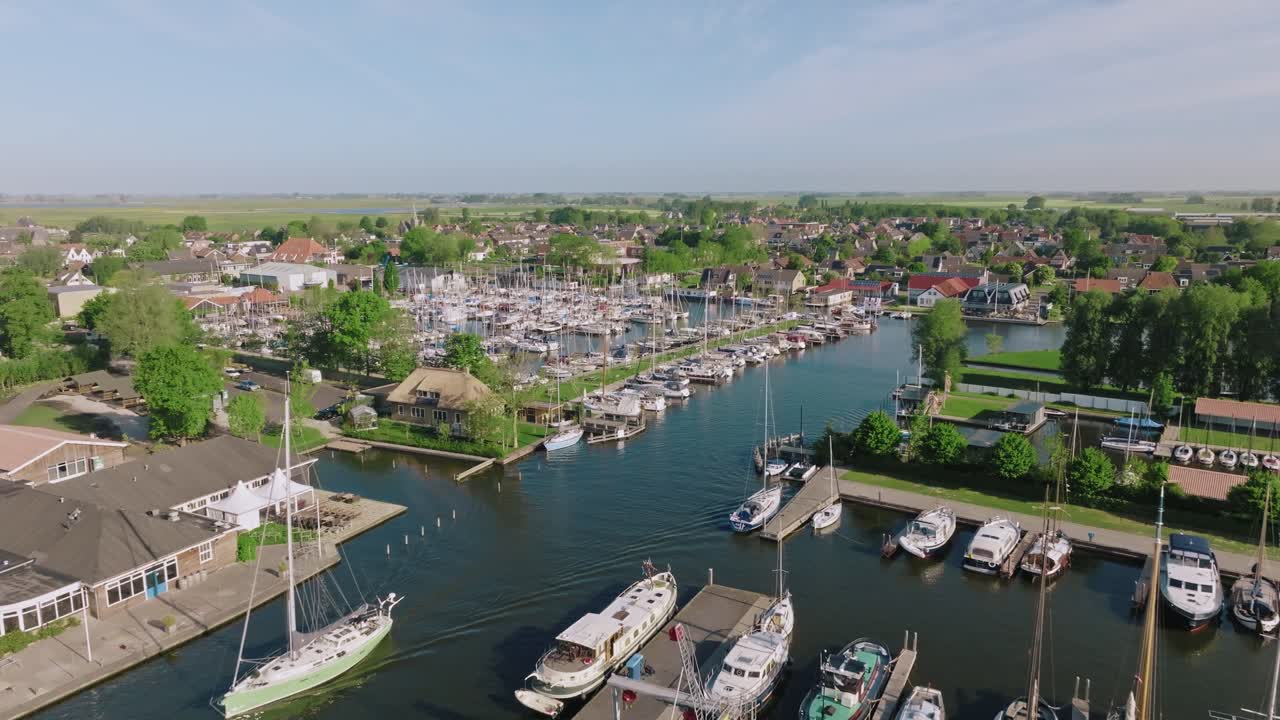 Drone slowly zooms in over dozens of moored sailboats in Heeg marina surrounded by buildings and trees