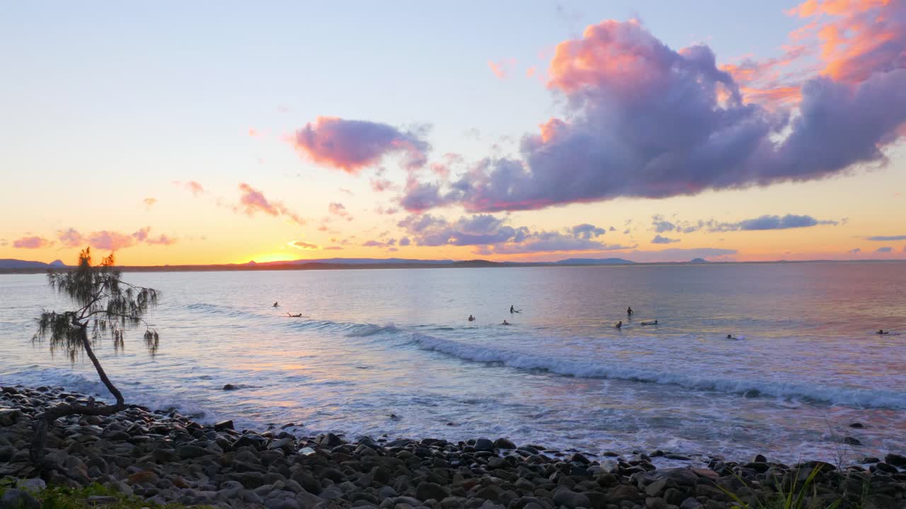 turistas surfeando en la playa ondulada con costa rocosa al atardecer en queensland