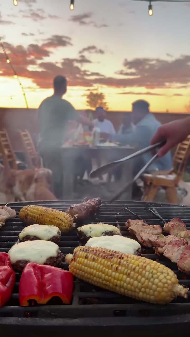 Video captures a low-angle view of a BBQ grill with friends dining at sunset in the background