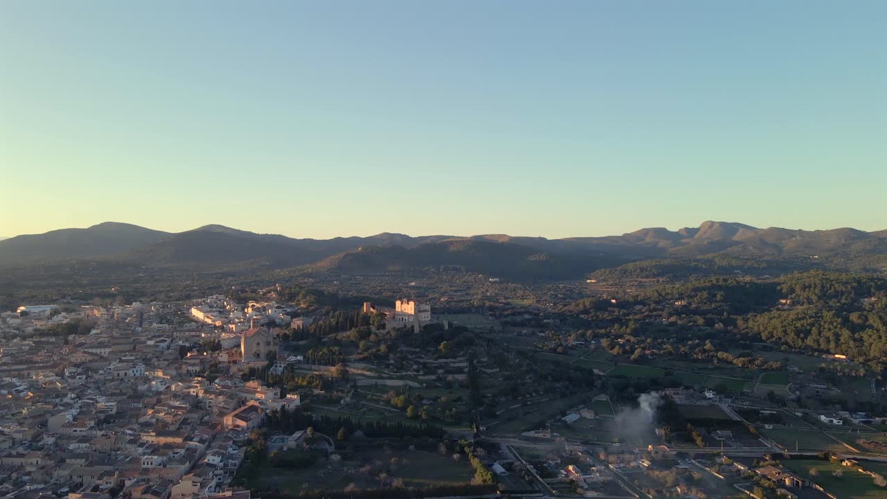 Experience breathtaking aerial views of Arta, Mallorca. The landscape features rolling hills, historic architecture, and serene natural beauty under the clear sky during sunset.