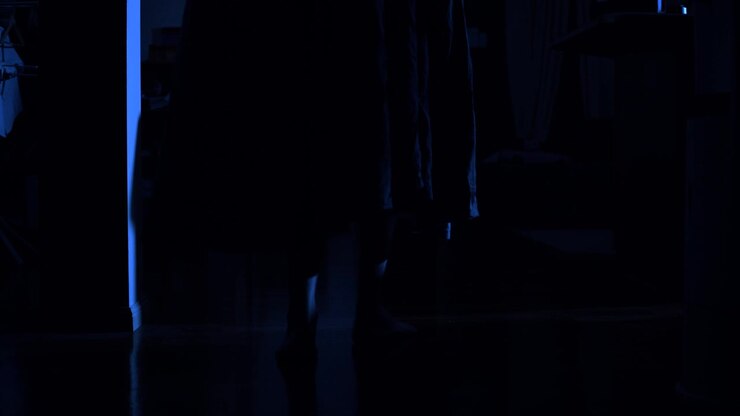 A dark, cloaked figure walks past camera towards a light source spilling from a hidden room at the dead of night in a chilly house