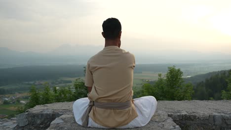 indian man doing hatha yoga meditation nadhi shuddhi at the edge of a stone castle wall in the morning sun at sunrise overlooking the valley bellow with fields and woods