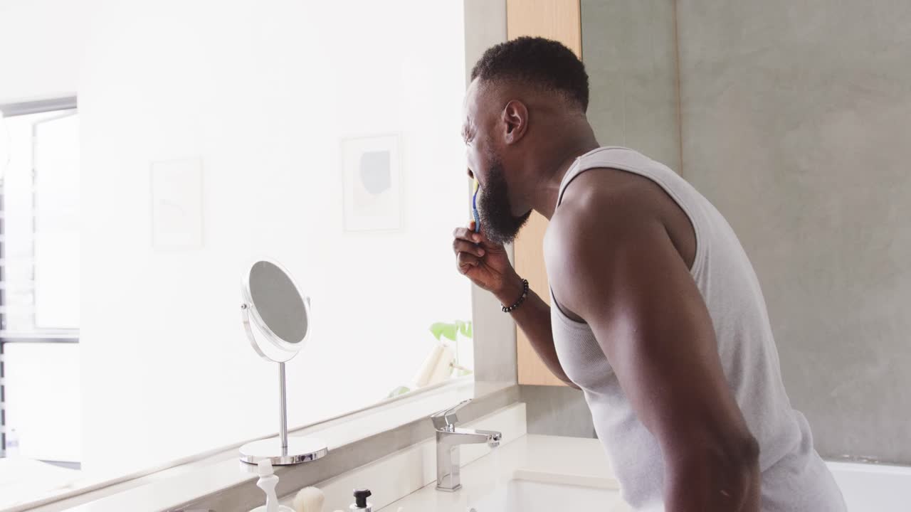 Uomo in canottiera bianca che si lava i denti al lavandino del bagno, guardandosi allo specchio con uno spazzolino blu.