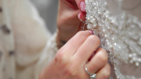 Close-up-Modedesigner-Für-Bräute-In-Seinem-Studio-Steckt-Nadeln-Spitzenhochzeitskleid-Fest.-Näherin-Kreiert-Ein-Exklusives-Hochzeitskleid.-Den-Umriss-Mit-Stecknadeln-Befestigen.-Kleines-Privatunternehmen.-Nähen-Sie-Strasssteine-Und-Kristalle-Mit-Faden-Und-Nadel-An-Das-Kleid.-Schmuckarbeiten.