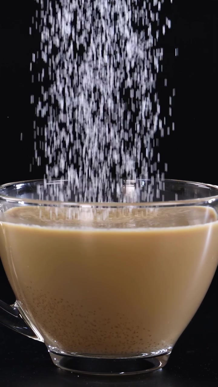 Granelli di zucchero che cadono a cascata in una tazza di caffè di vetro, su uno sfondo scuro, creando un effetto visivo dinamico