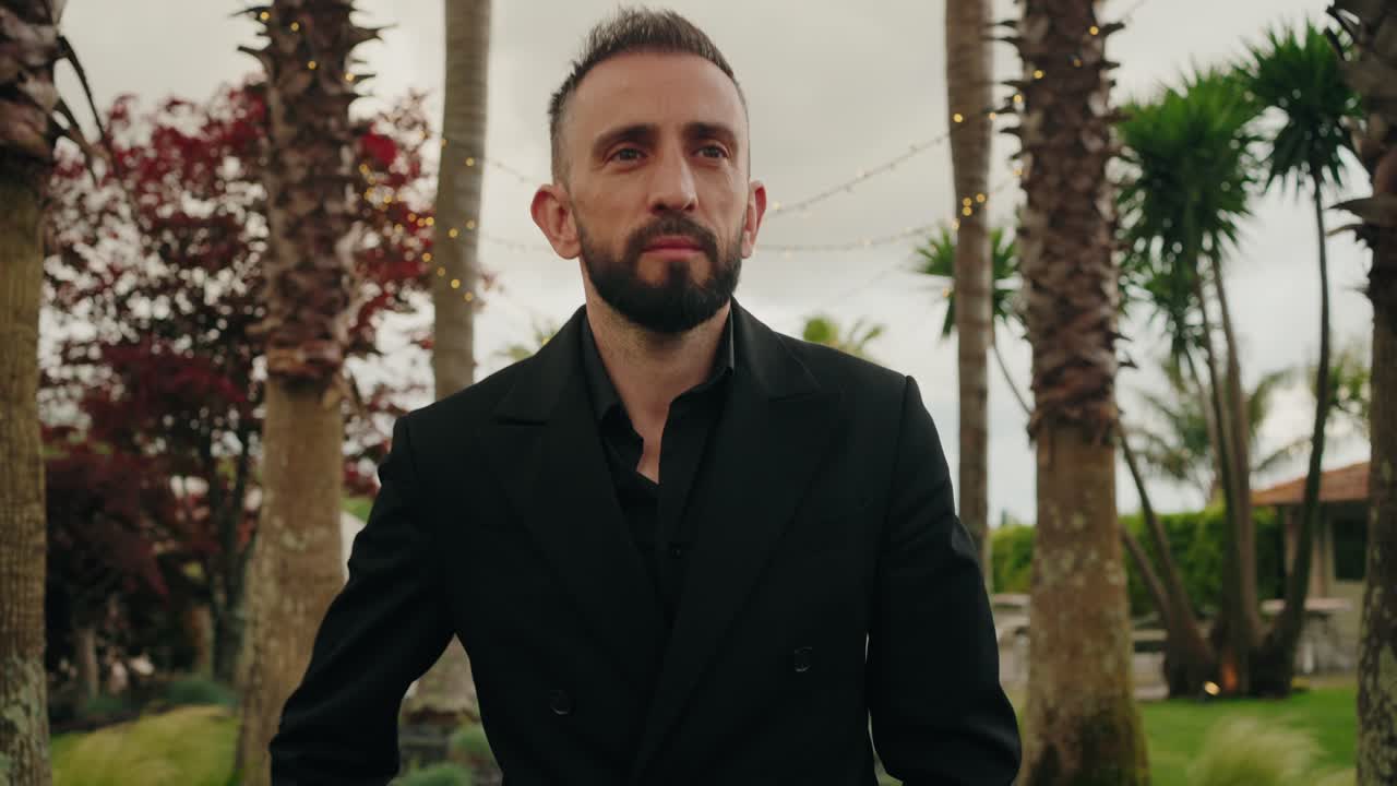 man in black suit walking outdoors with calm expression among palm trees