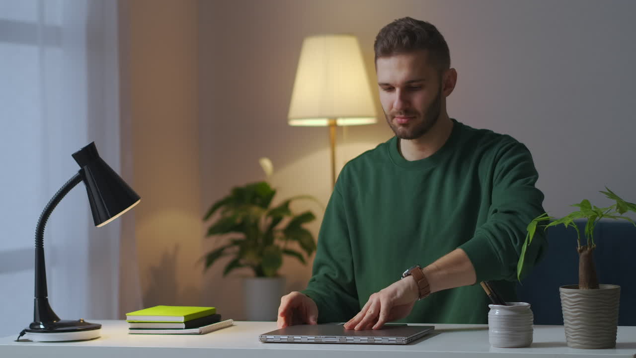 giovane uomo bello sta accendendo la lampada da tavolo e aprendo il portatile seduto nel soggiorno del suo appartamento alla sera lavoro remoto da casa e apprendimento online