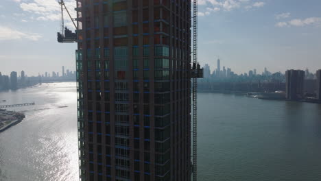 Pull-back-shot-of-construction-of-new-modern-tall-skyscraper-on-riverbank.-Construction-lift-moving-down-around-wall.-Downtown-skyscrapers-in-distance.-New-York-City,-USA