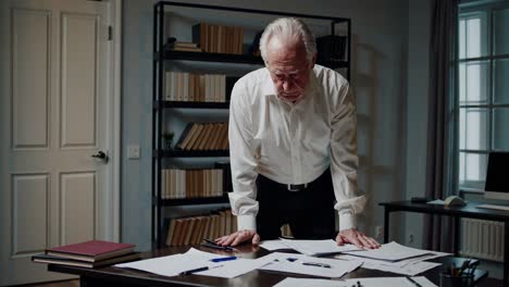 professional businessman analyzing paperwork, sitting tensely near bookshelf, displaying intense concentration while managing urgent corporate project under dim office lighting