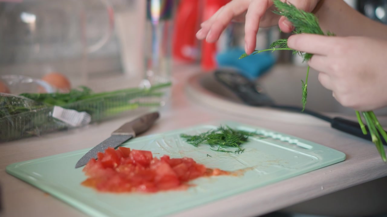 Preparazione di verdure in cucina. Preparazione degli ingredienti per l'insalata su un tagliere verde. Dettaglio del taglio di aneto fresco e pomodori a cubetti. Primo piano di una mano che strappa l'aneto e taglia i pomodori sul tagliere da cucina.