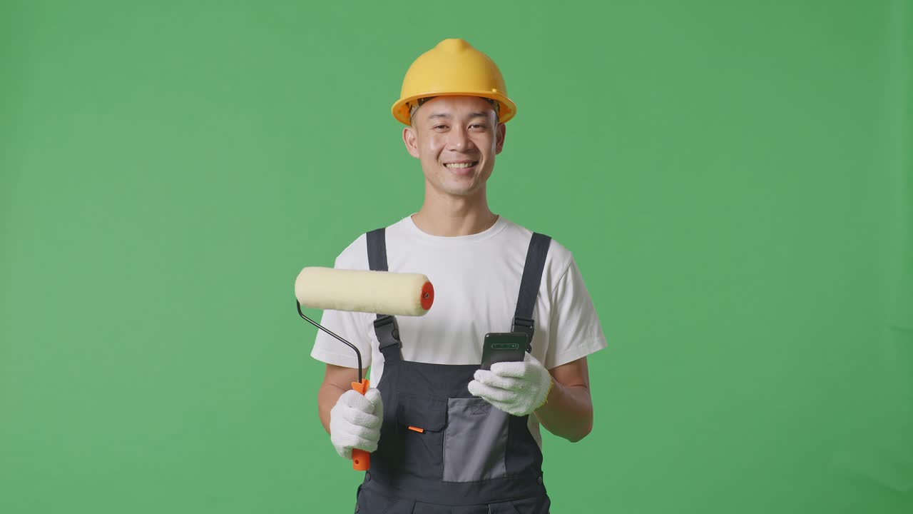 Asian Man Painter Wearing Safety Helmet Smiling And Using Smartphone While Standing In The Green Screen Background Studio