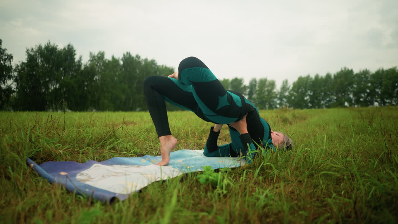 mujer acostada en una alfombra de yoga practicando la postura del puente con las manos apoyando su cintura en un campo de hierba bajo un cielo nublado, rodeada de naturaleza, los árboles están alineados en la distancia