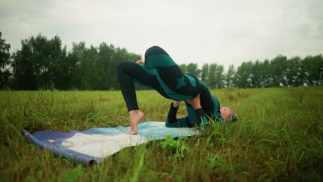 Woman lying on yoga mat practicing bridge pose with hands supporting her waist in grassy field under cloudy skies, surrounded by nature, trees are lined in the distance