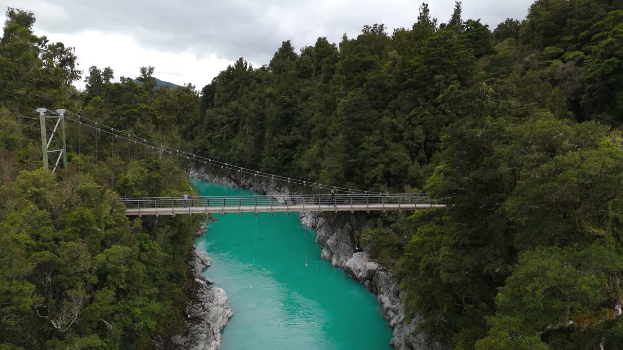 Drone pushes towards, girl crossing bridge over Hokitika River, flanked by vibrant green trees on both sides under cloudy sky in New Zealand.