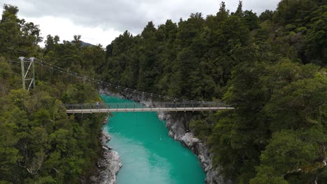 drone pushes towards, girl crossing bridge over hokitika river, flanked by vibrant green trees on both sides under cloudy sky in new zealand.