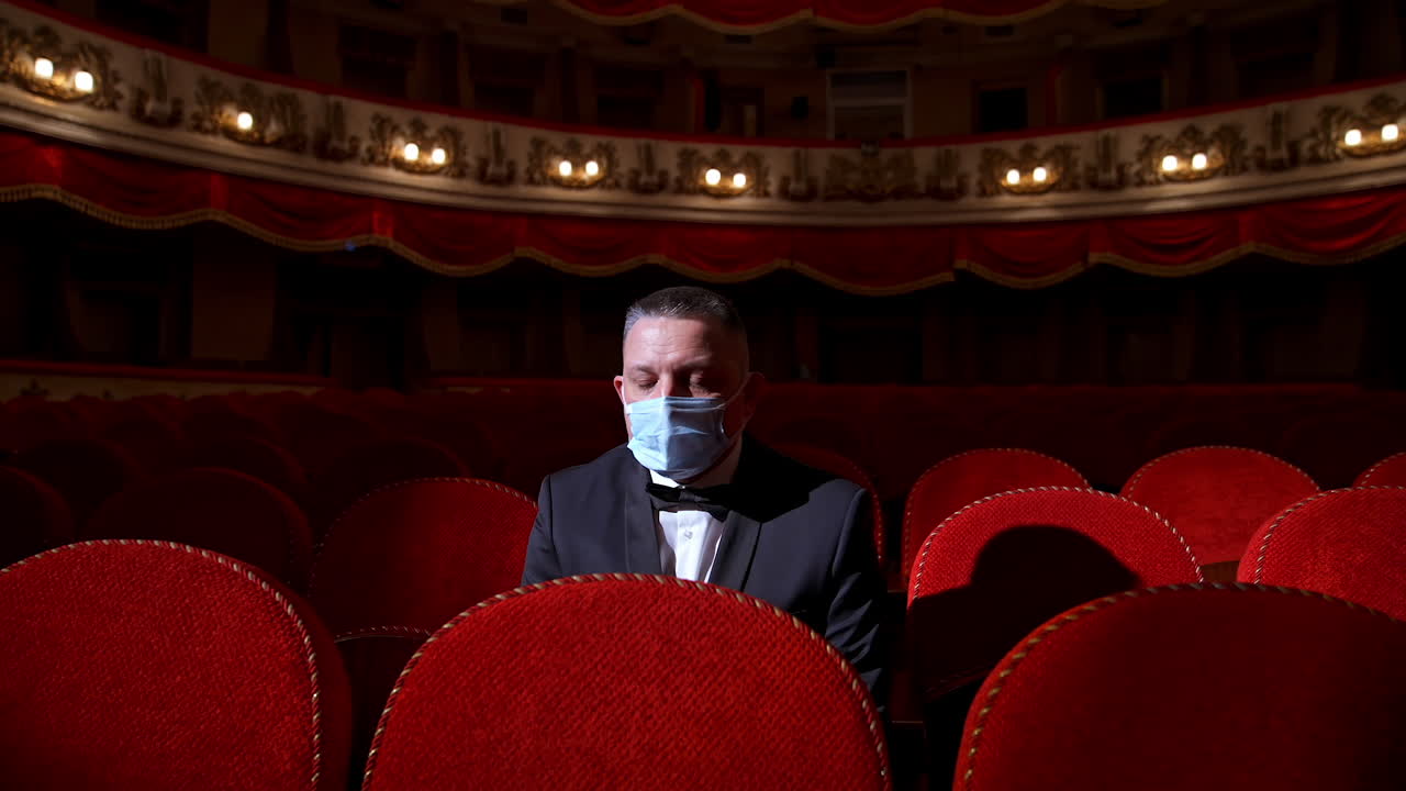 Visitor in suit and medical mask sitting in comfortable chairs alone on dark theater background. Male among rows of empty red chairs. Pandemic concept.