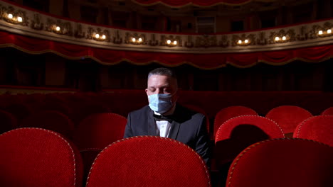 visitor in suit and medical mask sitting in comfortable chairs alone on dark theater background. male among rows of empty red chairs. pandemic concept.