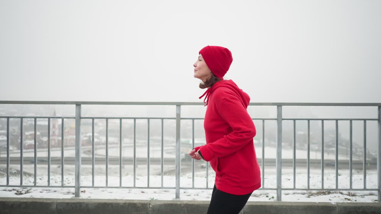 vista lateral de una mujer de negocios corriendo con gorra roja y chaqueta a lo largo de un camino nevado cerca de una barandilla de hierro, con vistas al paisaje urbano con puente y coches que pasan por debajo en una atmósfera de invierno niebla