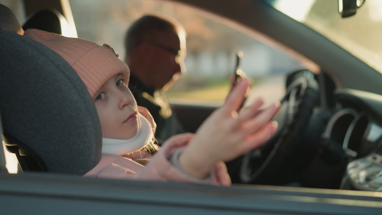 una joven, con un sombrero de punto rosa y una bufanda blanca, se sienta pacíficamente en un coche, agitando suavemente la mano mientras mira por la ventana. un hombre está ligeramente borroso, sosteniendo un teléfono mientras está sentado en la parte delantera